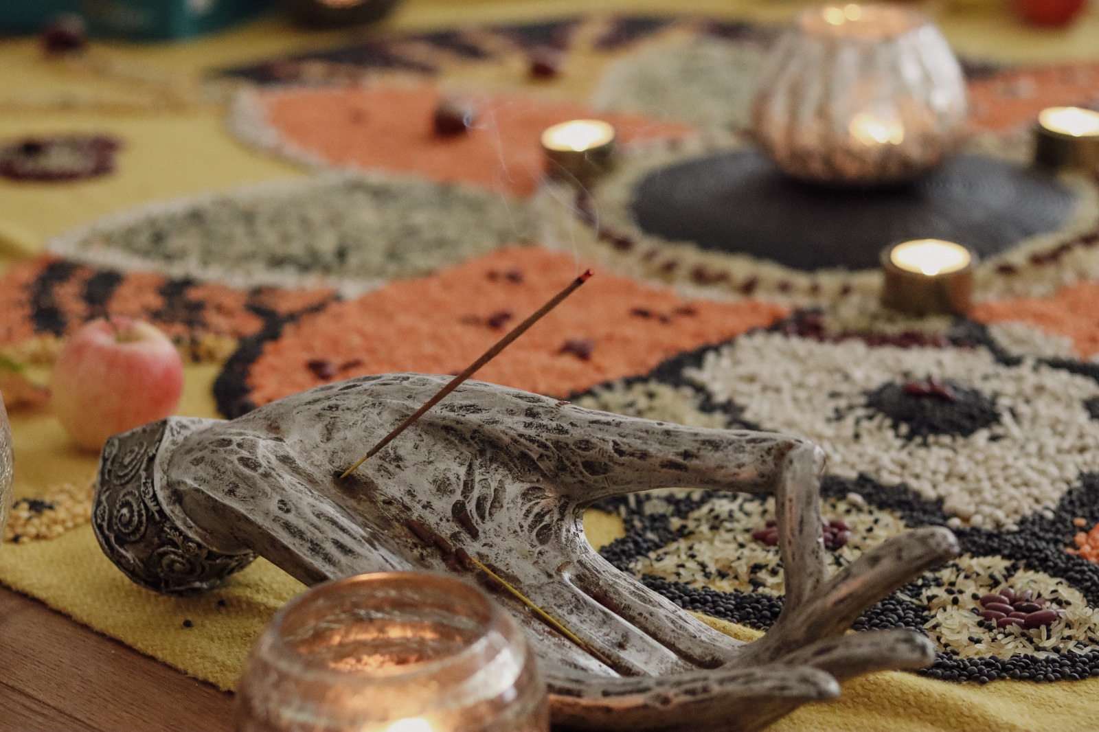 Bild mit einer dekorativen Handfigur aus hellem Material. In den Fingern steckt ein brennendes Räucherstäbchen, aus dem eine feine Rauchfahne nach oben steigt. Um die Hand herum stehen mehrere kleine Teelichter, deren warmes Licht die Umgebung weich ausleuchtet. Alles befindet sich auf einem gelben Tuch. Auf dem Tuch liegt ein kunstvoll gelegtes Mandala aus verschiedenen Körnern und Hülsenfrüchten.